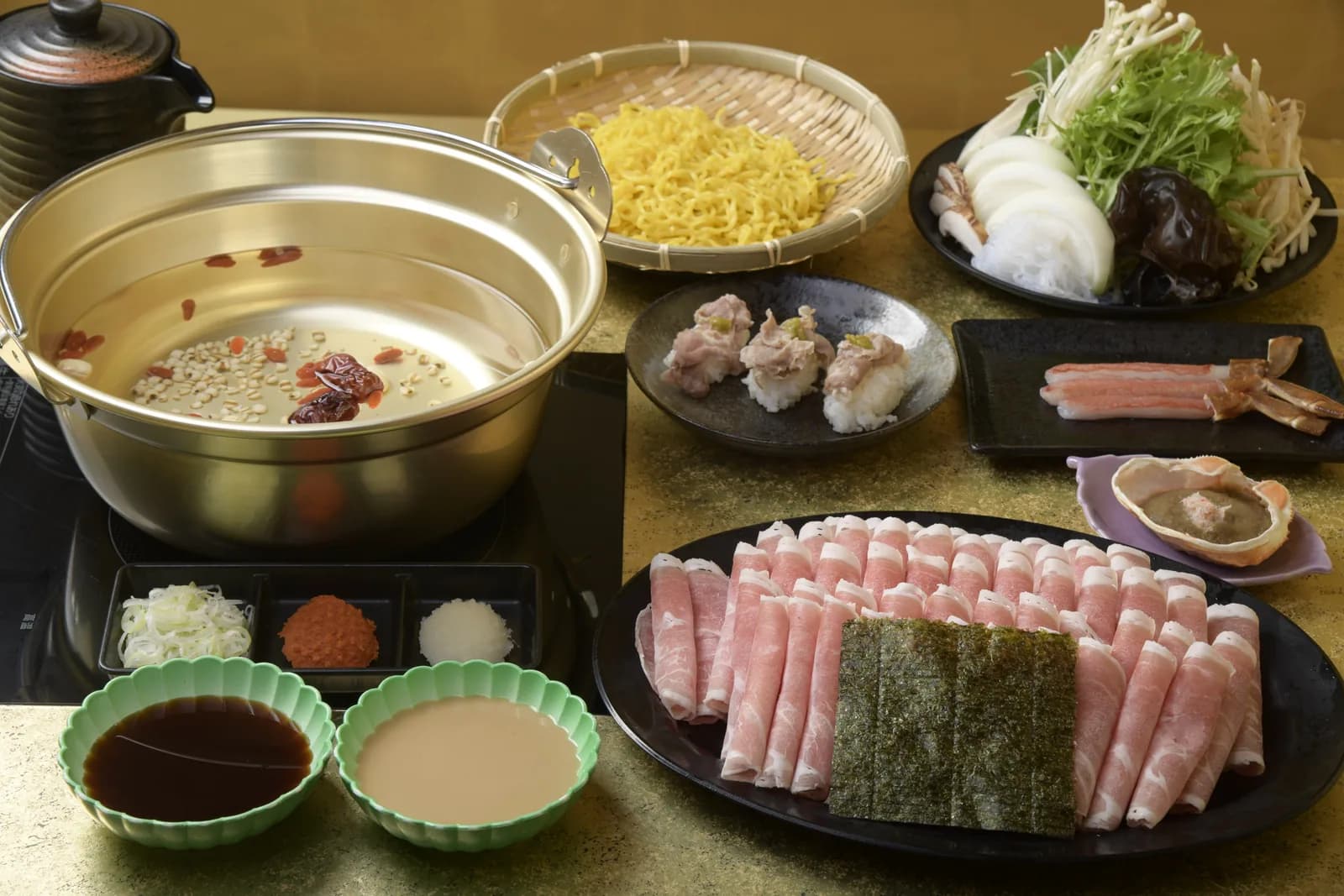 金色の鍋を据えた卓の全景。黒い大皿にロール状のラム肉を並べ、たまご麺・野菜・薬味・つみれを配する