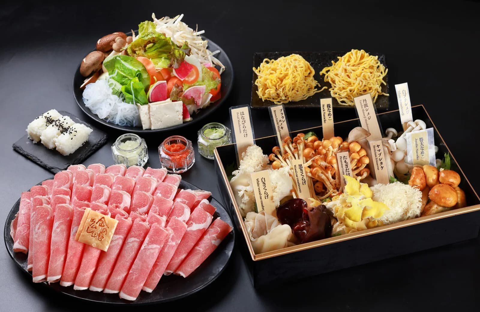 黒い卓を真上から。木札を立てたきのこの重箱、薄切りラム肉の大皿、サラダ、たまご麺二玉、おにぎり、薬味瓶が並ぶ