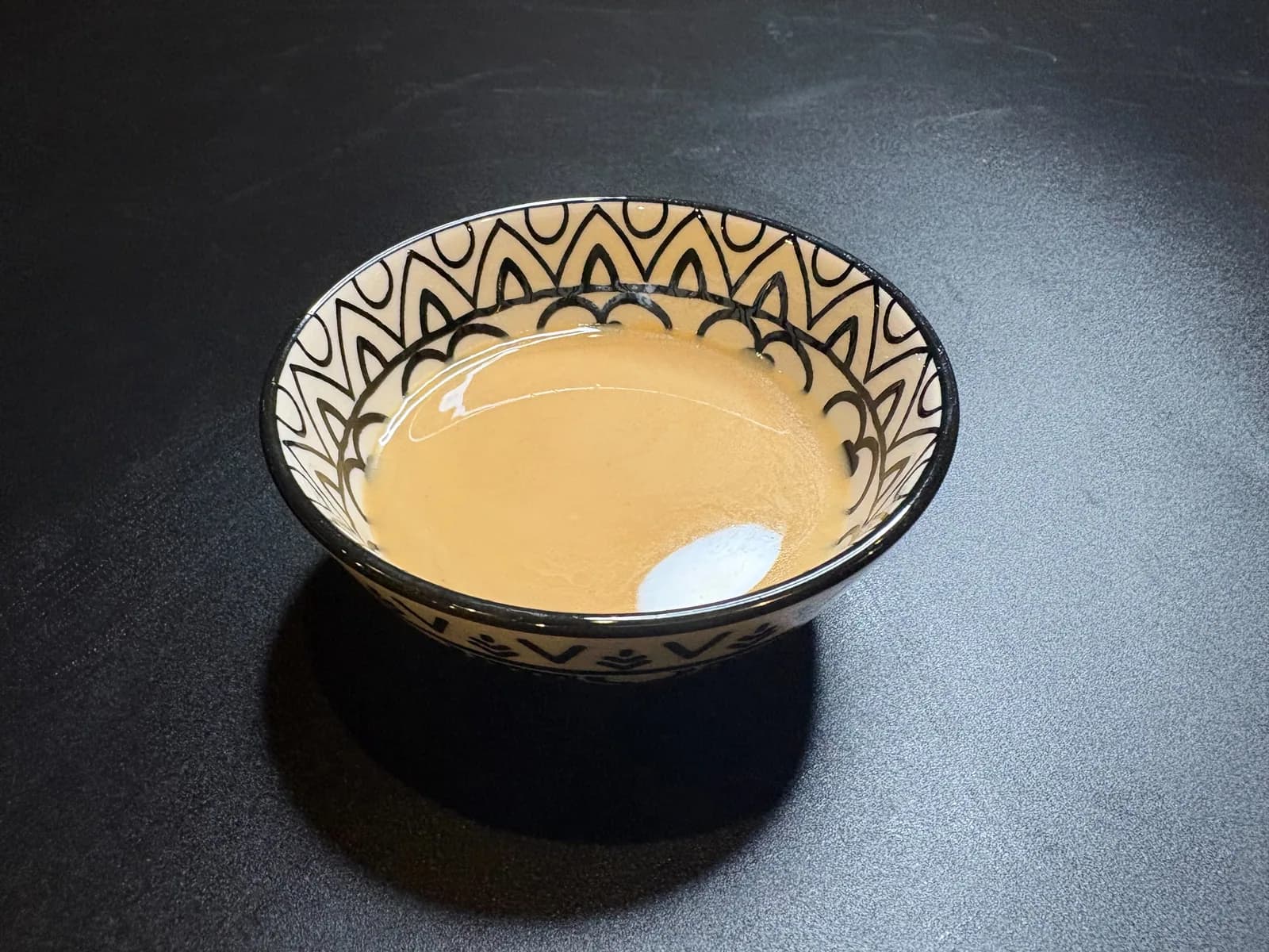 A small bowl with a black geometric pattern on white, set on a black table and filled with thick, milky white sesame sauce