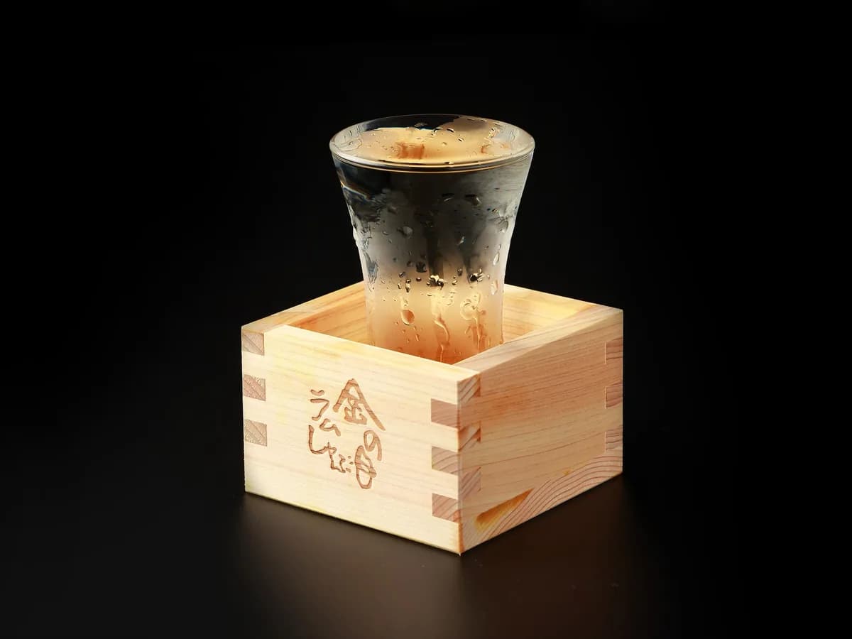 Chilled sake poured to the brim into a glass set in a plain wood masu branded KINNOME Lamb Shabu; droplets glisten against a black background