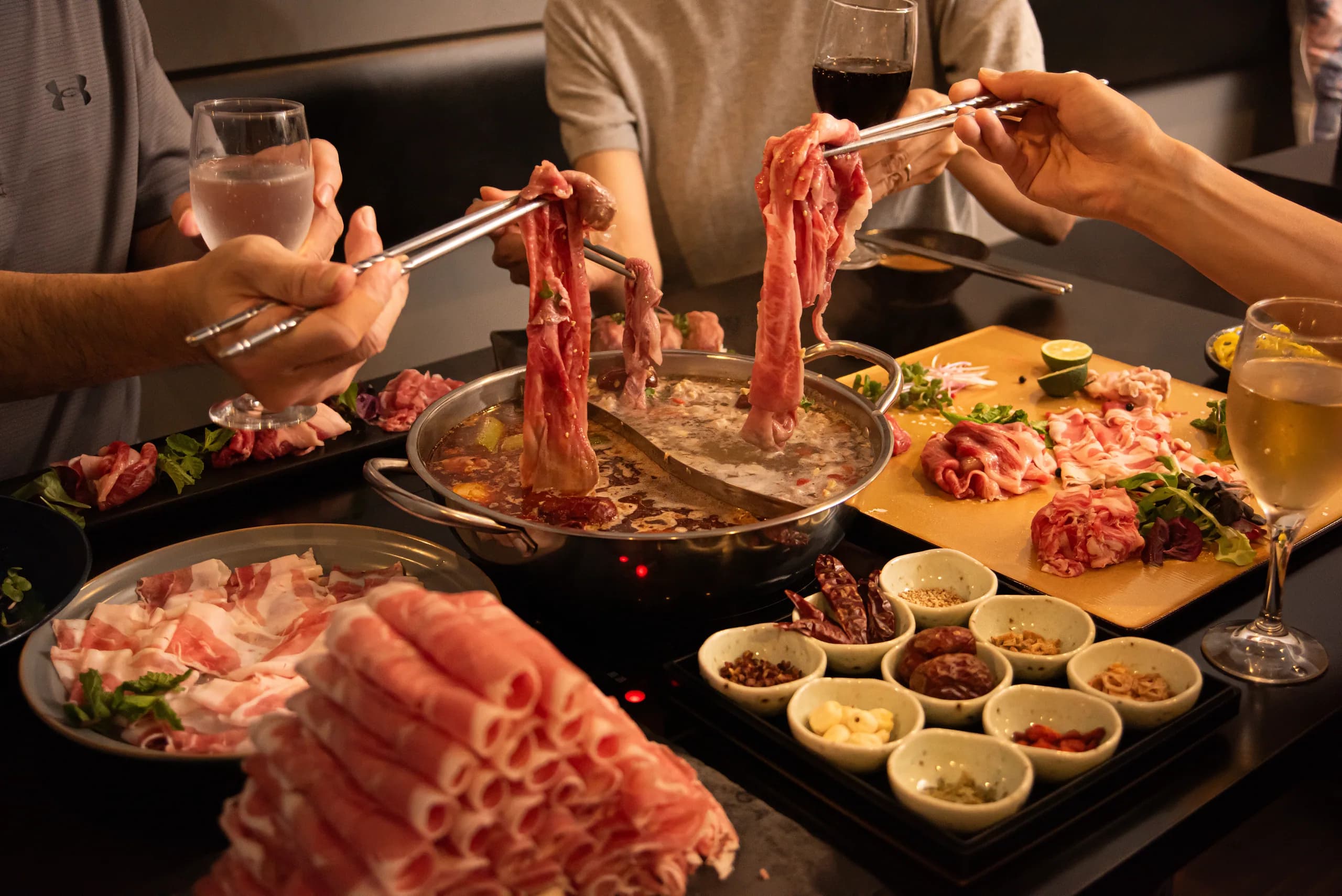 二人が箸で薄切りラム肉を二色鍋から引き上げる瞬間。手前にロールラムの山、金の板に盛った肉と薬味九種が並ぶ