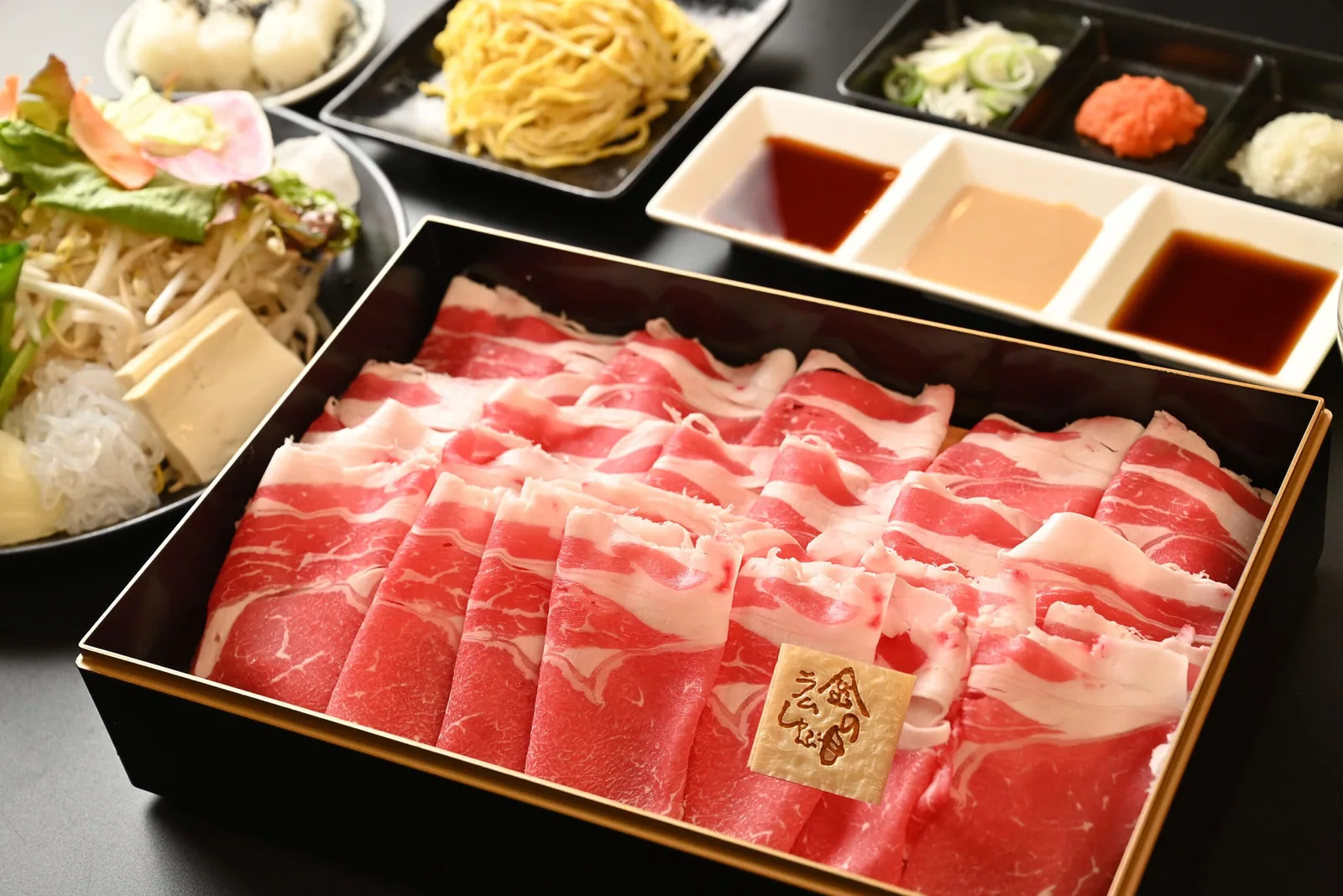 黒背景の卓上。金縁の重箱を埋め尽くす薄切りラム肉に木札が載り、サラダ、たまご麺、タレ三種と薬味三種が控える