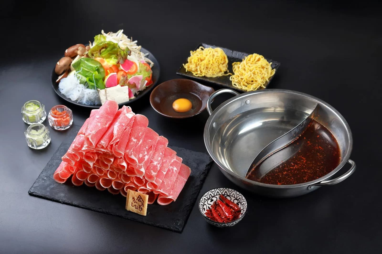 黒背景の卓上俯瞰。麻辣と白湯の二色鍋、ロール状のラム肉を敷いたスレート、サラダ、たまご麺、生卵、薬味が並ぶ