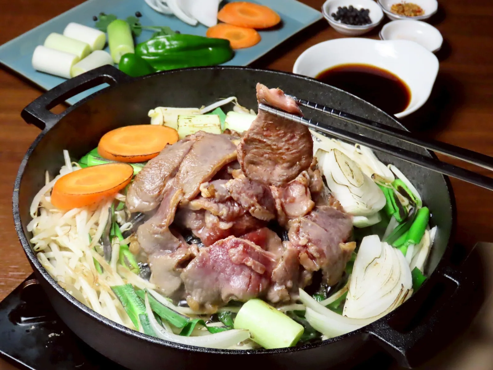 铁锅烤制的成吉思汗烤羊肉。用筷子夹起烤上色的羊肉与豆芽、大葱、洋葱、胡萝卜,后方是蘸料与佐料碟