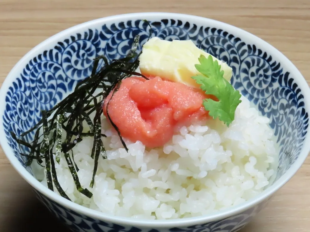 蓝色和风纹样碗中盛白饭,铺上明太子、海苔丝与黄油的收尾明太子饭。置于木纹餐桌上