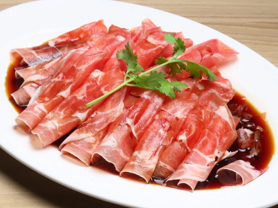 白い皿に薄切りラム肉を山形に盛り、黒酢だれをまわしかけてパクチーを添えた冷菜。木目のテーブルに置かれている