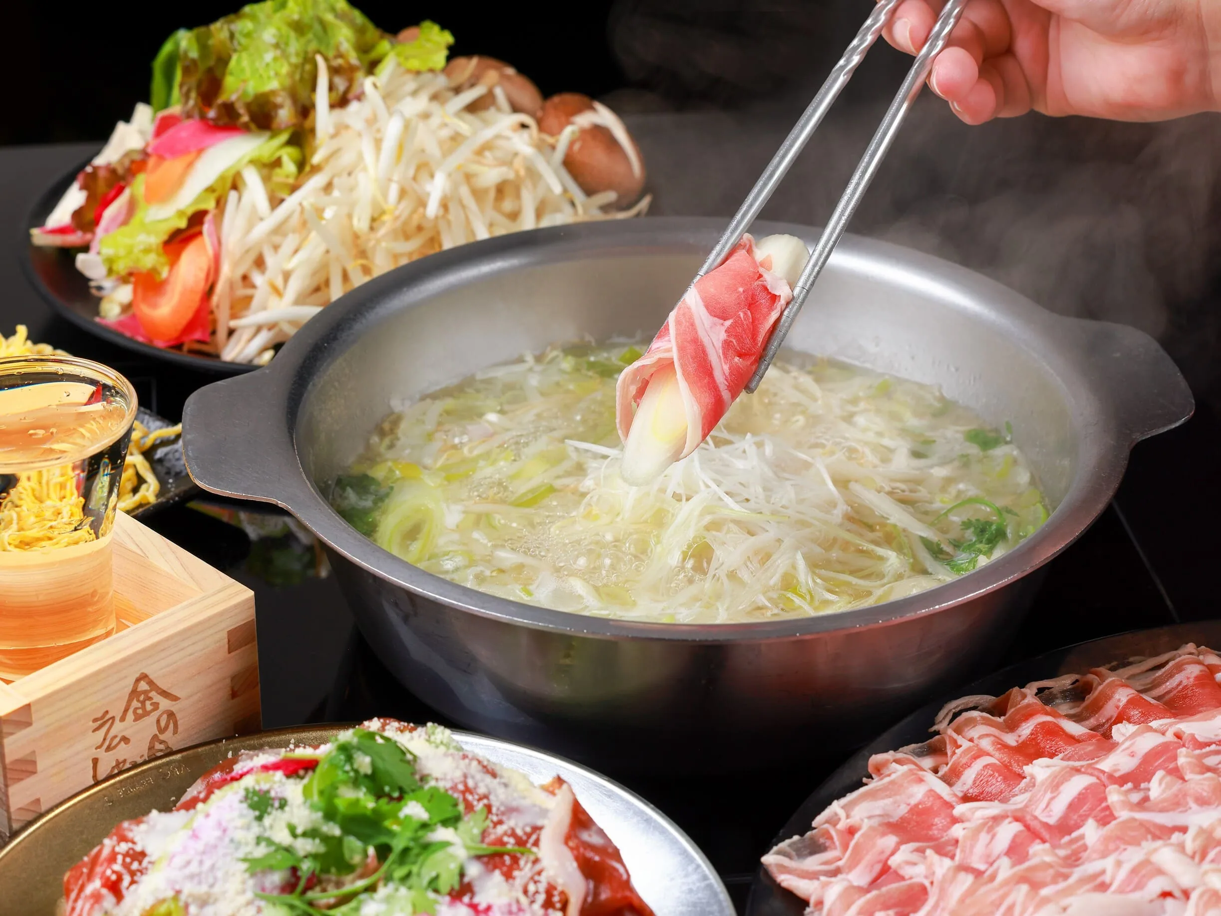 トングでつまんだ薄切りラム肉を白湯鍋に入れる瞬間。湯気の左に「金の目」の焼印が入った升のたまご麺が見える
